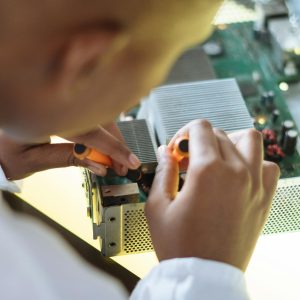 Crop of ethnic system administrator in uniform using screwdrivers and checking motherboard on electronic equipment during work in repair department